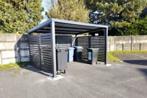 Abri en aluminium sur mesure EUROFACTORY avec brise-vues ajourés pour collectivités et espaces extérieurs, il abrite des poubelles.