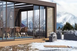 Pergola LINEA autoportée en aluminium sur mesure marron avec panneaux vitrés, située en montagne avec vue sur les sommets enneigés.